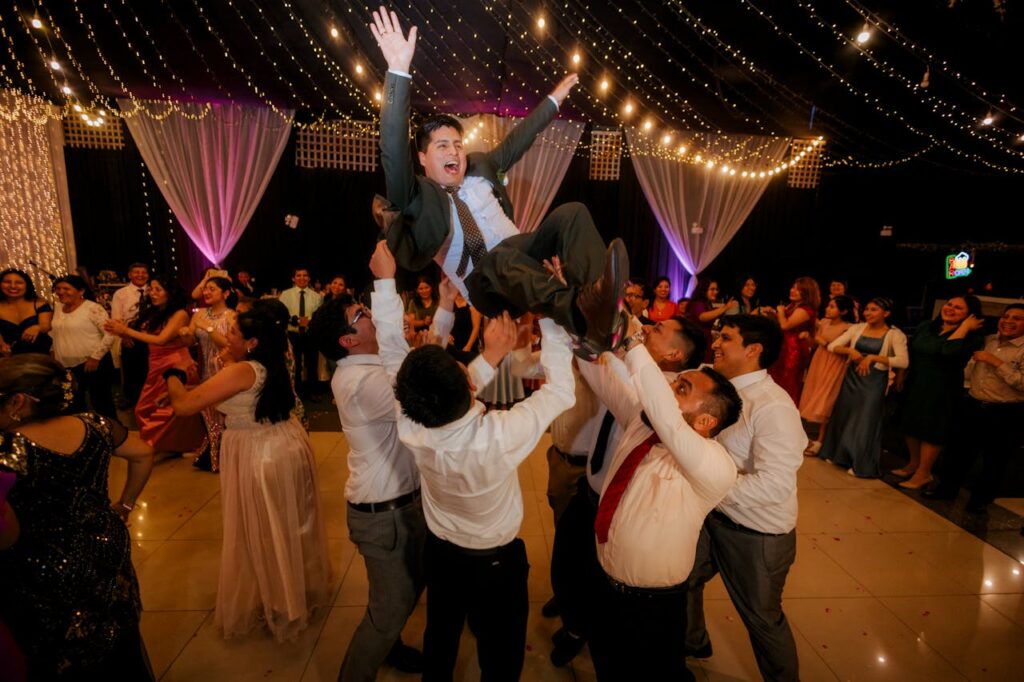 Guests celebrate joyfully at a wedding, lifting the groom amid laughter and dancing.
